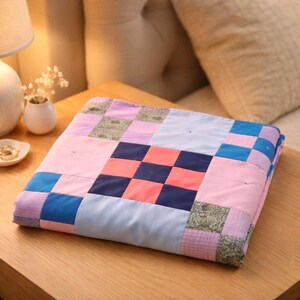 May include: A folded patchwork quilt featuring a mix of square patterns in pink, blue, purple, and green. The quilt is displayed on a wooden table, highlighting its geometric design and soft textures. The quilt is approximately 70 cm x 70 cm.
