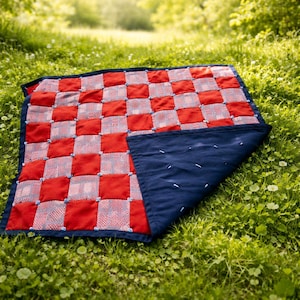 Op de afbeelding: Een rood-wit geblokte quilt met een marineblauwe achterkant, liggend op groen gras. De quilt heeft een rasterpatroon van rode vierkanten en witgepatroneerde vierkanten, met blauwe accenten.
