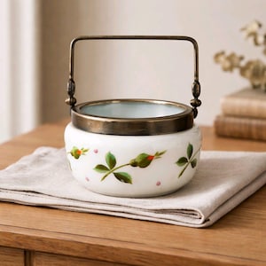 Puede incluir: Un recipiente de cerámica blanca con borde y asa de latón, decorado con hojas verdes, capullos rojos y puntos rosas. El recipiente descansa sobre un paño beige doblado, lo que sugiere un uso decorativo o de servicio.