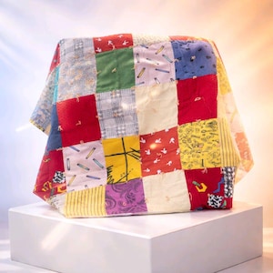Op de afbeelding: Een kleurrijke patchworkquilt gedrapeerd over een witte kubus. De quilt heeft een verscheidenheid aan stoffen vierkanten in rood, groen, geel, blauw en patronen. De quilt is een mix van texturen en patronen, wat een levendige en gezellige uitstraling creëert.