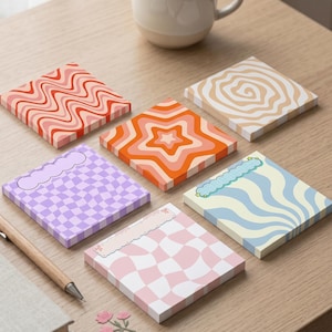 May include: Six square notepads with retro designs in various colours, including orange, pink, and blue. The pads feature patterns such as wavy lines, stars, and checkerboards. A pen and a mug are also visible on the wooden surface.