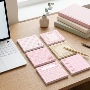 Puede incluir: Una colección de notas adhesivas rosas estampadas con varios diseños, incluyendo lazos, marcos y un gato, dispuestas sobre un escritorio de madera. También hay un portátil, un bolígrafo, unas tijeras y libros.