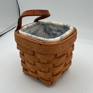 Peut inclure: Un panier carré en bois tressé avec une anse en cuir marron. Le panier est doté d'une doublure en tissu clair avec un motif botanique bleu de feuilles et de baies. L'extérieur est composé de bandes de bois superposées fixées par des clous.