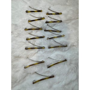 May include: A collection of hair barrettes with a tortoiseshell pattern in shades of brown and yellow. The barrettes have a silver-colored metal clasp. The hair accessories are arranged on a white, textured surface.