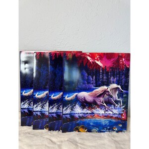 May include: Four notebooks with a fantasy art design. The covers feature two white horses running through water, with a mountain range and forest in the background. The color palette includes shades of blue, purple, and red.
