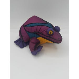 May include: A plush frog toy constructed from fabric panels in shades of purple, teal, and yellow. The stuffed animal features prominent round eyes with yellow irises and black pupils, along with webbed feet and stitched detailing throughout.