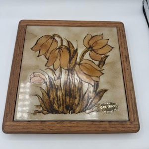 Peut inclure: Un dessous de plat carré composé d'un carreau en céramique orné d'une illustration florale dans les tons marron et beige, encadré de bois. Le carreau présente des fleurs tombantes et un autocollant ovale marqué S.R.G. OAK TRIVET THE DALLES, OR.