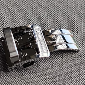 Könnte beinhalten: Nahaufnahme einer silbernen Uhrenschließe und eines Armbands. Die Schließe ist rechteckig mit dem eingravierten Text "Grand Seiko" und "Seiko". Das Armband ist gebogen und trägt ebenfalls den Text "Grand Seiko". Die Artikel befinden sich auf einer strukturierten grauen Oberfläche.