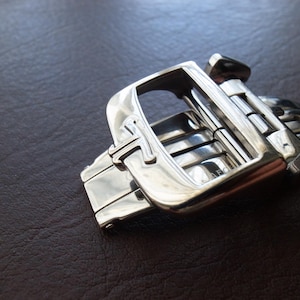 May include: Close-up of a shiny, silver watch clasp. The clasp is made of multiple interlocking pieces and has a rectangular shape with rounded edges. The metal reflects light, creating highlights and shadows. The background is a dark, textured surface.