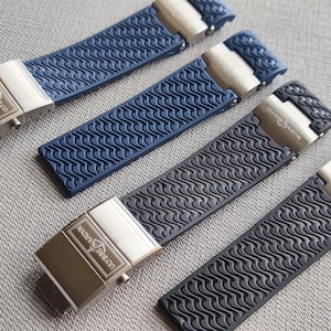 May include: Four watch straps in blue and black with a wave pattern. Each strap has a silver clasp with the text "ULYSSE NARDIN" engraved on it. The straps are made of a flexible material.