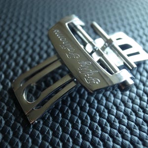 May include: A close-up of a silver metal watch buckle with the engraved text "Fifty Fathoms". The buckle is composed of two interlocking parts with a rectangular shape and a polished finish. The background is a textured, dark blue surface.