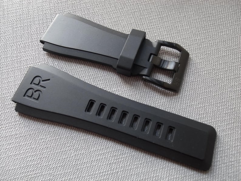 以下が含まれることがあります： バックルとストラップに「BR」の文字がエンボス加工された黒い時計ストラップ。 ストラップには一連の長方形の切り欠きがあります。 バックルも黒です。