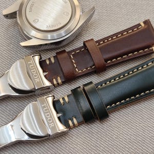 May include: Two leather watch straps in brown and dark green with cream stitching. The straps have silver clasps with the brand name "TUDOR" engraved. A watch case back is visible in the background.