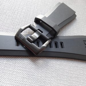 以下が含まれることがあります： 黒い腕時計のストラップとそれに合うバックル。ストラップは柔軟な素材で作られており、端には「BR」の文字が入っています。バックルはマットブラック色で、ストラップを固定するように設計されています。