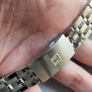 Puede incluir: Una correa de reloj de acero inoxidable en tono plateado con un cierre desplegable de metal cepillado. El centro del cierre muestra una letra T en relieve con un acabado texturizado. La correa consta de eslabones pulidos y articulados.