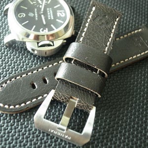 May include: A dark brown textured leather watch strap with white contrast stitching and a silver-toned metal buckle engraved with the word Panerai, positioned next to a silver-cased wristwatch with a black dial labelled Luminor Marina Panerai.