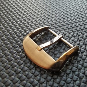 以下が含まれることがあります： 曲線的なデザインのゴールドトーンの金属製腕時計バックル。 バックルは、ピンと丸みを帯びたエッジを備えた長方形のフレームです。 背景は、テクスチャ加工された濃い灰色の表面です。