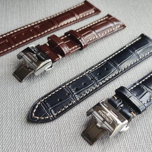May include: Close-up of three leather watch straps in brown and black. The straps have a crocodile pattern with white stitching. Two straps are brown, and one is black. The straps have silver clasps with the "Longines" logo.