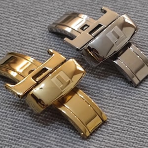 Może przedstawiać: Dwie metalowe klamry zatrzaskowe do zegarka, jedna w kolorze polerowanego złota, a druga w kolorze polerowanego srebra. Każda klamra posiada mechanizm zawiasowy z centralną prostokątną płytką z wytłoczonym logo w kształcie litery T.