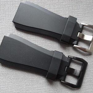 以下が含まれることがあります： シルバーと黒の金属バックルが付いた2つの黒い時計ストラップ。 ストラップはマットな素材でできており、端が角度になっています。 バックルは長方形で、クラシックなデザインです。