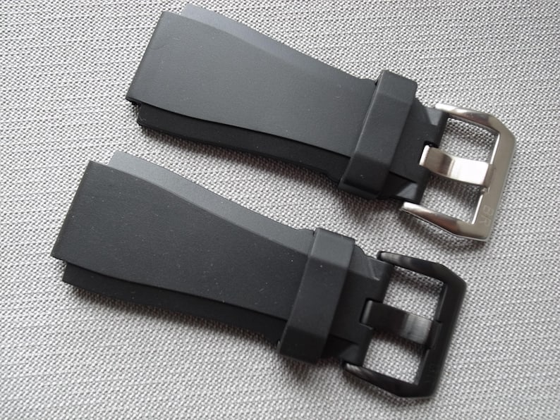 以下が含まれることがあります： シルバーと黒の金属バックルが付いた2つの黒い時計ストラップ。 ストラップはマットな素材でできており、端が角度になっています。 バックルは長方形で、クラシックなデザインです。