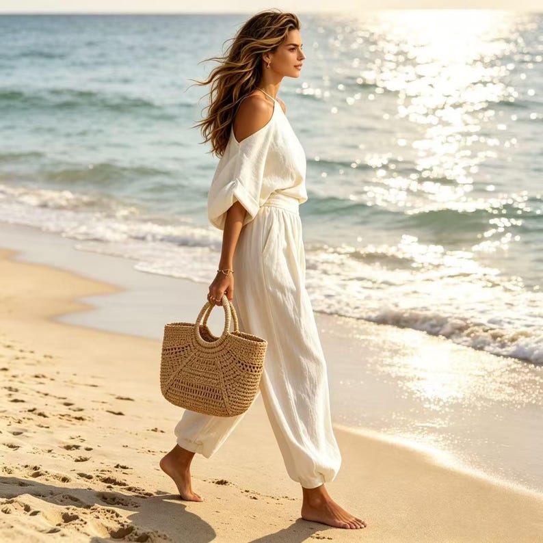 K&ouml;nnte beinhalten: Eine Person geht an einem Sandstrand entlang und tr&auml;gt eine geflochtene Stroh-Tragetasche. Sie tr&auml;gt ein wei&szlig;es Oberteil mit freier Schulter und eine passende Hose. Das Meer und die Sonne sind im Hintergrund und schaffen eine sommerliche Szene.