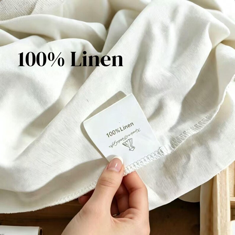 K&ouml;nnte beinhalten: Nahaufnahme von wei&szlig;em Leinenstoff mit dem Text "100% Linen" in fetten schwarzen Buchstaben. Ein Etikett mit dem gleichen Text und einem Logo wird in der N&auml;he des Stoffes gehalten. Der Stoff wirkt weich und leicht zerknittert.