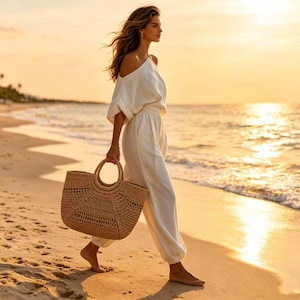 K&ouml;nnte beinhalten: Eine Person geht am Strand bei Sonnenuntergang, tr&auml;gt ein wei&szlig;es schulterfreies Oberteil und eine passende Hose. Sie tr&auml;gt eine gewebte, hellbraune Tragetasche mit runden Griffen. Die Tasche hat ein H&auml;keldesign. Der Hintergrund zeigt das Meer und einen goldenen Himmel.