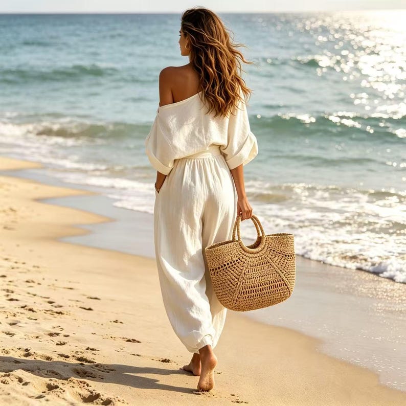 K&ouml;nnte beinhalten: Eine Person in einem wei&szlig;en schulterfreien Oberteil und passender Hose geht an einem Sandstrand entlang und tr&auml;gt eine gewebte Stroh-Tragetasche. Das Meer ist im Hintergrund zu sehen, mit sanft am Ufer pl&auml;tschernden Wellen. Das Outfit ist l&auml;ssig und strandtauglich.