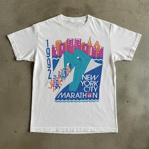 May include: White t-shirt featuring a colorful graphic for the 1992 New York City Marathon. The design includes a cityscape in shades of orange, pink, and purple, with the words "New York City Marathon" in blue. The shirt has short sleeves.