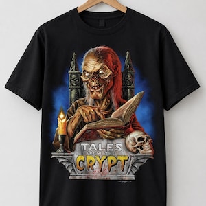 May include: A black short-sleeve t-shirt featuring a graphic print of the Crypt Keeper sitting on a throne, reading an open book. A lit candle and a human skull are visible on a stone base with the text Tales From The Crypt printed in yellow.
