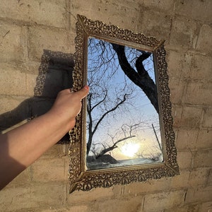 May include: A gold-toned ornate mirror with a detailed, intricate frame reflects a view of a tree silhouetted against a blue sky with a bright sun shining through the branches.