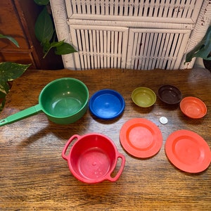 Könnte beinhalten: Ein Set aus vintage Plastik-Spielzeug-Küchenutensilien und Geschirr. Das Set enthält einen grünen Topf mit Griff, eine rote Schüssel mit zwei Griffen, einen blauen Teller, einen grünen Teller, eine braune Schüssel, eine weiße Schüssel und zwei orangefarbene Teller.