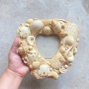 Op de afbeelding: Een hartvormig fotolijstje gemaakt van schelpen. De lijst is gemaakt van verschillende soorten schelpen, waaronder zanddollars, schelpen en sint-jakobsschelpen. De lijst is lichtbeige van kleur.