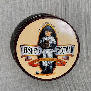 May include: Round Hershey's Chocolate tin with a baseball theme. The tin features an illustration of a young baseball player in a vintage uniform, holding a glove. The text reads "Hershey's Chocolate" and "The Great American Chocolate Bar."