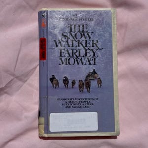 May include: A hardcover book titled "The Snow Walker" by Farley Mowat. The cover is a light blue with the title in large white letters. The book features an illustration of dogs pulling a sled.