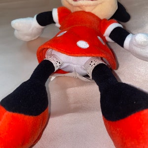 Op de afbeelding: Een rood en zwart pluche speelgoed van Minnie Mouse, die een rode jurk met witte stippen draagt. Het speelgoed heeft een wit gezicht met zwarte ogen en een zwarte neus. Het speelgoed ligt op zijn rug met zijn benen in de lucht.