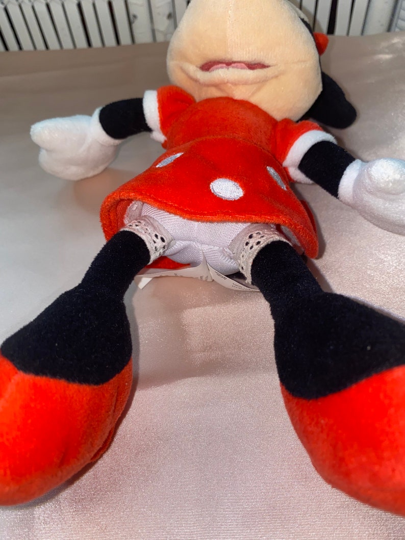 Op de afbeelding: Een rood en zwart pluche speelgoed van Minnie Mouse, die een rode jurk met witte stippen draagt. Het speelgoed heeft een wit gezicht met zwarte ogen en een zwarte neus. Het speelgoed ligt op zijn rug met zijn benen in de lucht.
