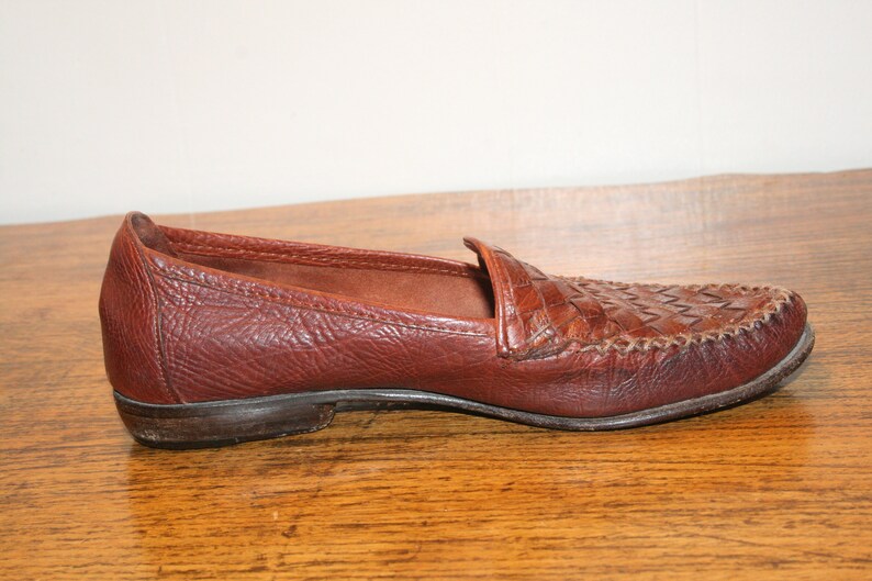 Op de afbeelding: Een paar bruine leren loafers met een geweven patroon op de neus. De loafers hebben een simpel ontwerp en een comfortabele uitstraling.