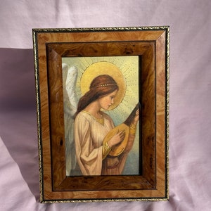 以下が含まれることがあります： リュートを弾く天使を描いた額縁付きの絵画。天使は翼、後光、そして長い流れるようなガウンを着ています。絵画は薄い青色の背景に描かれています。