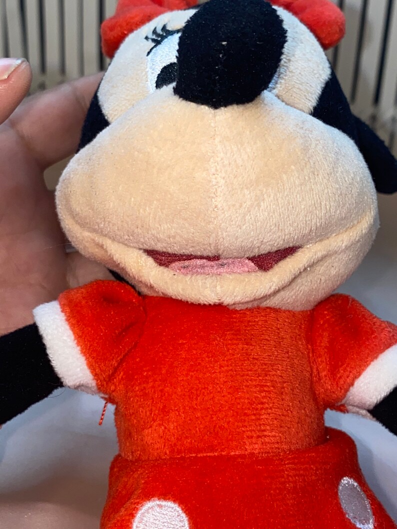 Op de afbeelding: Een pluche speelgoed van Minnie Mouse, die een rode jurk met witte stippen draagt. Ze heeft zwarte ogen, een zwarte neus en een rode strik op haar hoofd.
