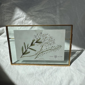 May include: A rectangular, gold-toned metal picture frame with a clear glass front. Inside is a botanical print with green leaves and small white dots. The frame is labeled "ISAAC JACOBS" and fits a 10cm x 15cm photo.