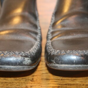 Puede incluir: Un par de mocasines de cuero negro con un detalle cosido alrededor del borde. Los zapatos tienen un aspecto ligeramente desgastado.