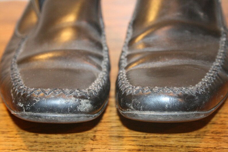 Puede incluir: Un par de mocasines de cuero negro con un detalle cosido alrededor del borde. Los zapatos tienen un aspecto ligeramente desgastado.