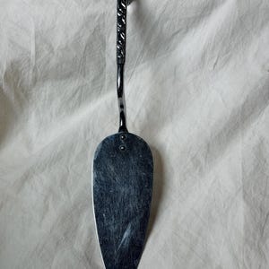 May include: A silver-colored cake server with a decorative handle. The handle has a textured pattern, and the blade is oval-shaped. The server is made of metal and appears to be handcrafted.