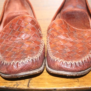 Op de afbeelding: Een paar bruine leren loafers met een geweven patroon. De loafers hebben een gestikte naad rond de rand van de zool.