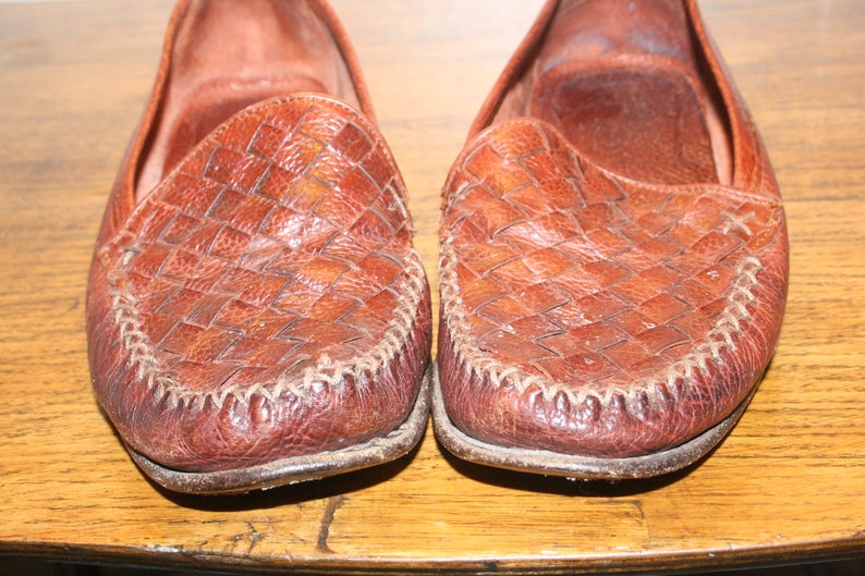 Op de afbeelding: Een paar bruine leren loafers met een geweven patroon. De loafers hebben een gestikte naad rond de rand van de zool.