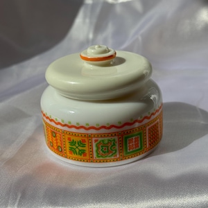 May include: A small, white ceramic jar with a lid. The jar features a decorative band of orange, yellow, and green geometric patterns around its base. The lid has a small, orange-trimmed knob.