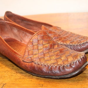 Op de afbeelding: Een paar bruine leren loafers met een geweven patroon. De loafers hebben een spitse neus en een platte hak.