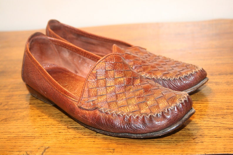 Op de afbeelding: Een paar bruine leren loafers met een geweven patroon. De loafers hebben een spitse neus en een platte hak.